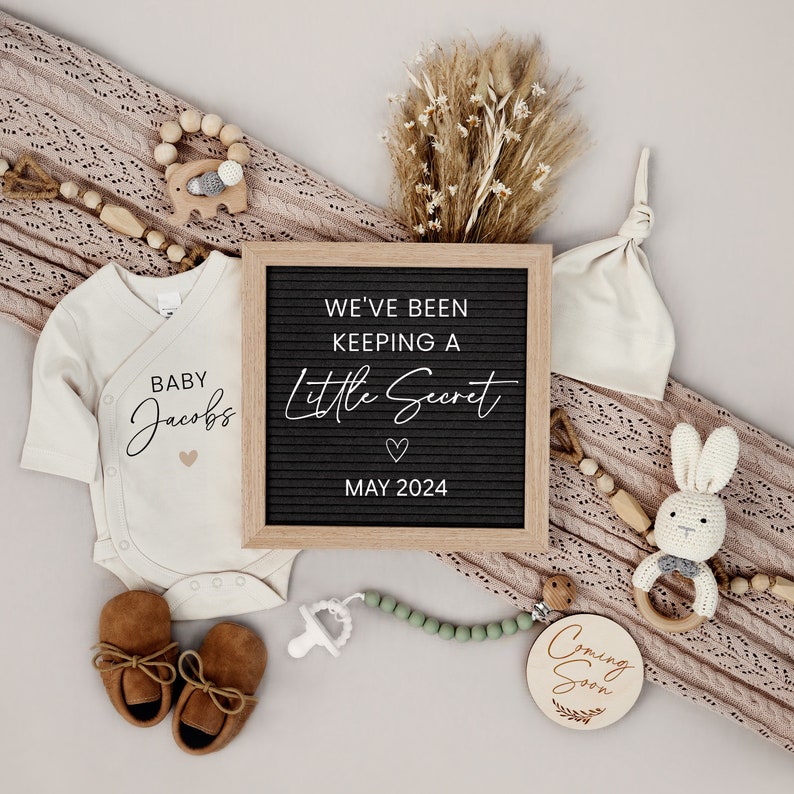 Puede incluir: Un anuncio de nacimiento con un tablero de letras que dice "We've been keeping a little secret May 2024". Hay un body blanco con el texto "Baby Jacobs" y un gorro de punto marr&oacute;n. El anuncio tambi&eacute;n incluye un letrero de madera que dice "Coming Soon" y un sonajero de madera con un conejito adjunto.