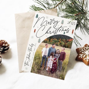 Pu&ograve; includere: Un biglietto di Natale con una foto di una famiglia in un campo. Il biglietto ha uno sfondo bianco con alberi di Natale verdi e rossi e fiocchi di neve. Il testo "Buon Natale 2023" &egrave; scritto in corsivo nero. La famiglia &egrave; in piedi in un campo con foglie autunnali sullo sfondo. Il biglietto dice "The Johnson Family" in basso.