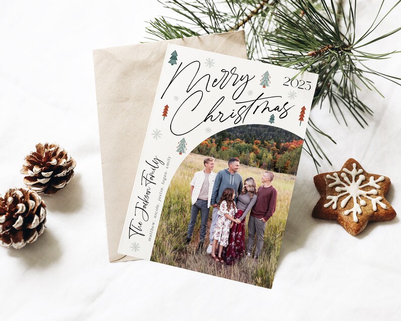 Pu&ograve; includere: Un biglietto di Natale con una foto di una famiglia in un campo. Il biglietto ha uno sfondo bianco con alberi di Natale verdi e rossi e fiocchi di neve. Il testo "Buon Natale 2023" &egrave; scritto in corsivo nero. La famiglia &egrave; in piedi in un campo con foglie autunnali sullo sfondo. Il biglietto dice "The Johnson Family" in basso.