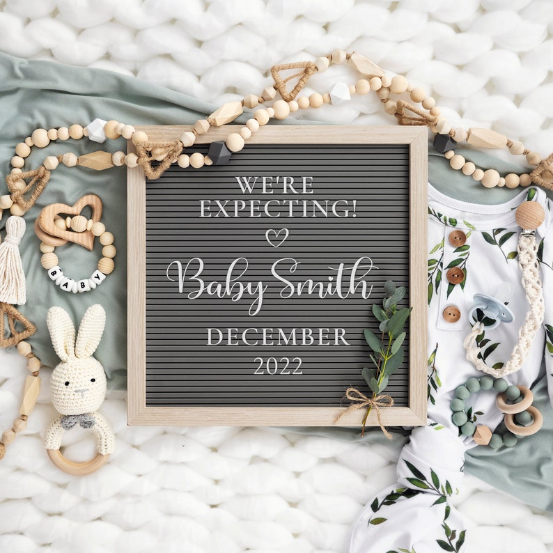 May include: A letter board with the text "We're Expecting! Baby Smith December 2022" surrounded by wooden beads, a crocheted bunny, and a baby outfit with a leafy pattern.