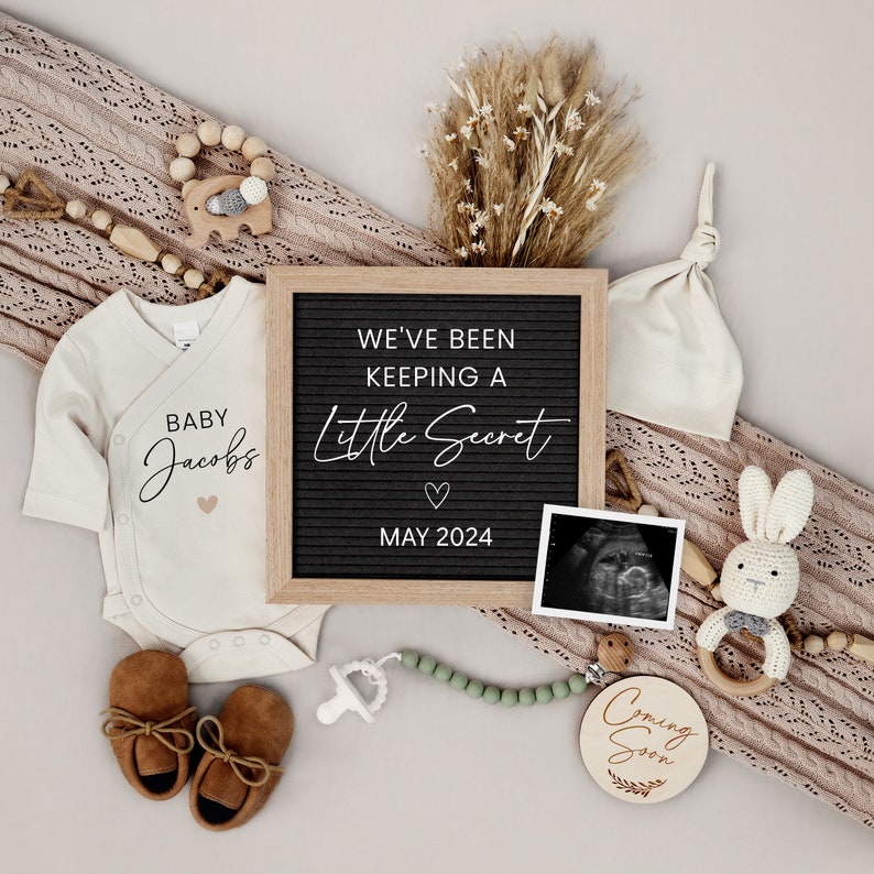 Puede incluir: Un anuncio de revelaci&oacute;n de g&eacute;nero con un body blanco que dice "Baby Jacobs" y un tablero de letras negro que dice "We've been keeping a little secret, May 2024". Hay un letrero de madera que dice "Coming Soon" y un gorro blanco de punto.