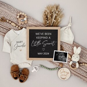 Puede incluir: Un anuncio de embarazo con un tablero de letras que dice "We've Been Keeping A Little Secret May 2024". El anuncio incluye un body de beb&eacute; que dice "Baby Jacobs", un par de zapatos de beb&eacute; marrones, un sonajero de madera, un c&iacute;rculo de madera que dice "Coming Soon", un chupete, un collar de cuentas, una manta de punto, un conejito de peluche, un gorro de beb&eacute; y una imagen de ultrasonido.