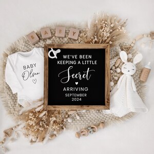 May include: A black chalkboard sign with white text announcing "We've been keeping a little secret, arriving September 2024". The sign is surrounded by a white onesie with the text "Baby Olsen" and other baby-themed decorations.