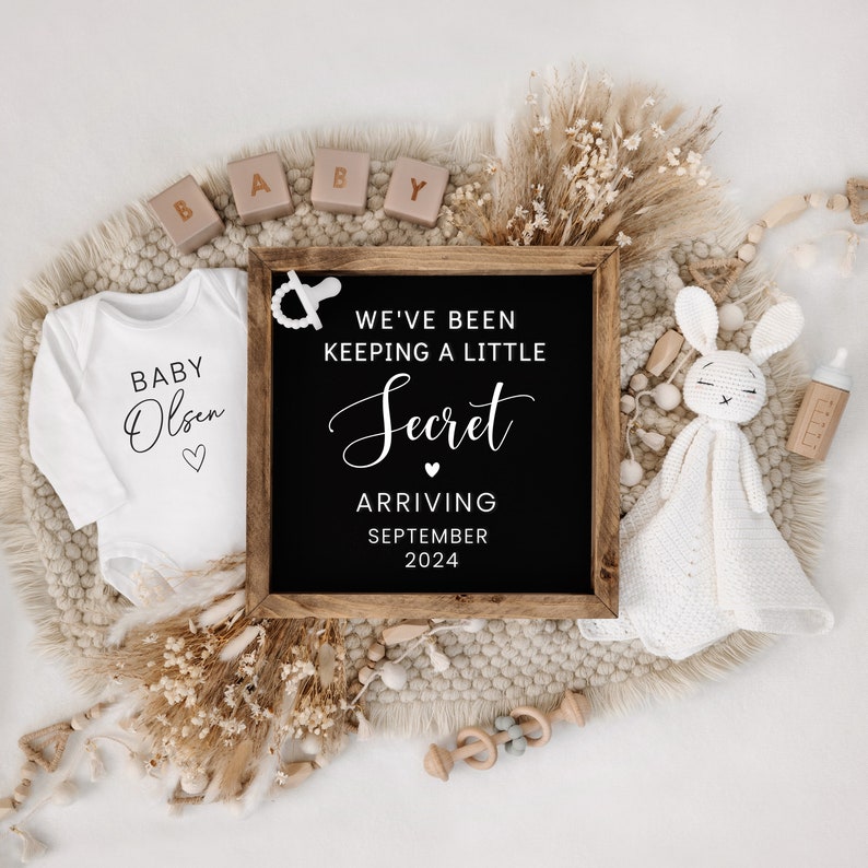 May include: A black chalkboard sign with white text announcing "We've been keeping a little secret, arriving September 2024". The sign is surrounded by a white onesie with the text "Baby Olsen" and other baby-themed decorations.