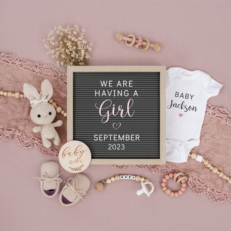 Op de afbeelding: Een roze en witte gender reveal-aankondiging met een letterbord waarop staat "We are having a girl September 2023". Er is een baby-romper met de tekst "Baby Jackson" met een roze hartje, een houten babyratel, een houten baby-speelgoed, een paar babyschoentjes, een houten bord met de tekst "Baby Girl", een gehaakt konijntje en een kralensnoer.