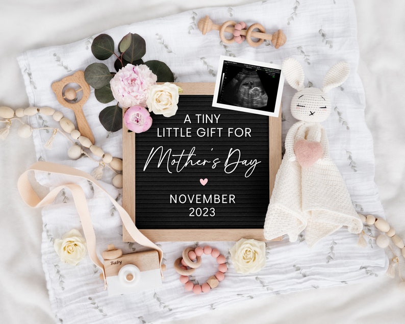 May include: A black letter board with the text "A tiny little gift for Mother's Day November 2023".  The board is surrounded by a white blanket, a crocheted bunny toy, a wooden camera, a wooden rattle, and pink and white flowers.