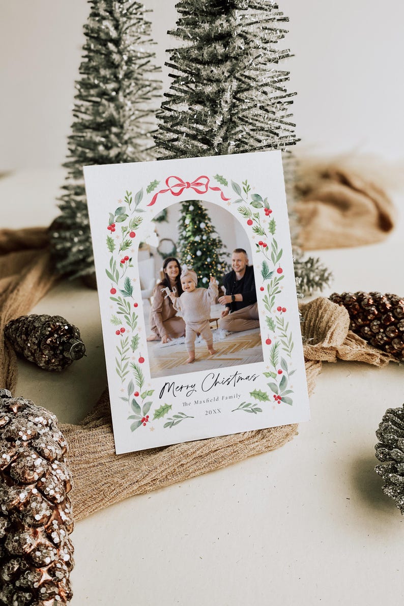 May include: A Christmas card with a watercolor wreath border featuring a red bow, holly, and greenery. A family photo is inside the wreath, showing a couple and their baby standing in front of a Christmas tree. The card reads "Merry Christmas The Maxfield Family 20XX".