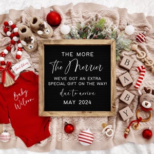 Puede incluir: Un mameluco rojo con el texto "Baby Wilson" en &eacute;l, rodeado de decoraciones navide&ntilde;as, incluyendo un letrero de pizarra que dice "The More The Merrier, We've Got An Extra Special Gift On The Way! Due To Arrive May 2024".