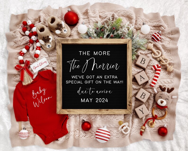 Puede incluir: Un mameluco rojo con el texto "Baby Wilson" en &eacute;l, rodeado de decoraciones navide&ntilde;as, incluyendo un letrero de pizarra que dice "The More The Merrier, We've Got An Extra Special Gift On The Way! Due To Arrive May 2024".