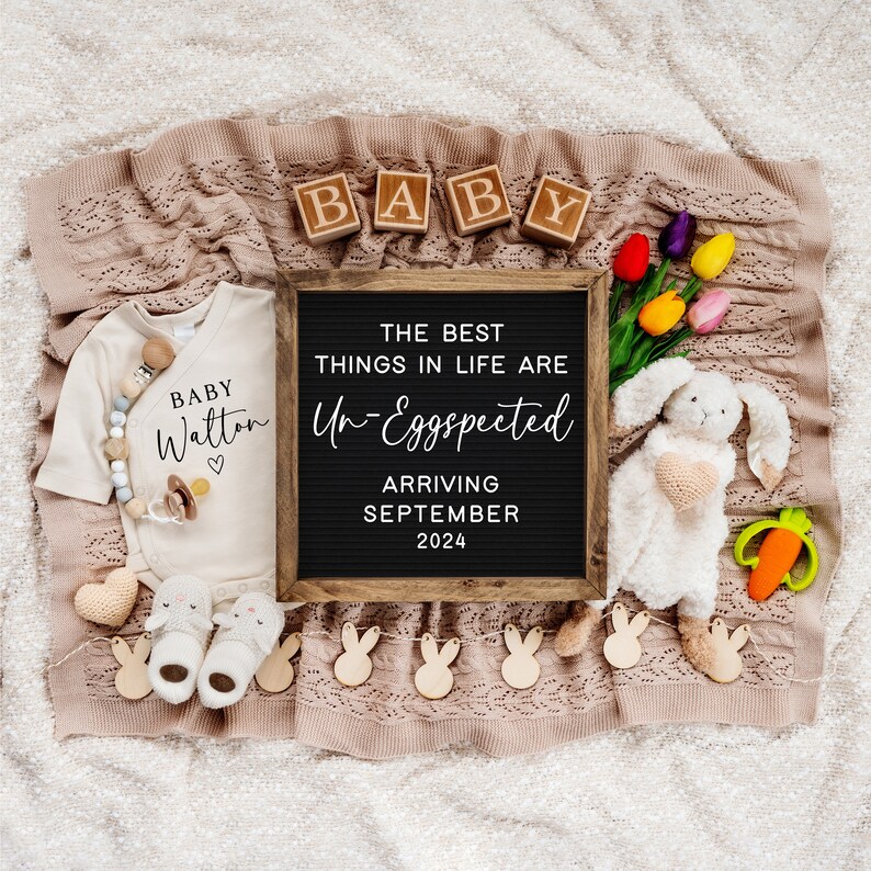 May include: A white onesie with the text "BABY Walton" and a heart. A letter board with the text "The best things in life are Un-Eggspected Arriving September 2024".  Wooden blocks spelling "BABY" and a white bunny stuffed animal with a crocheted heart.