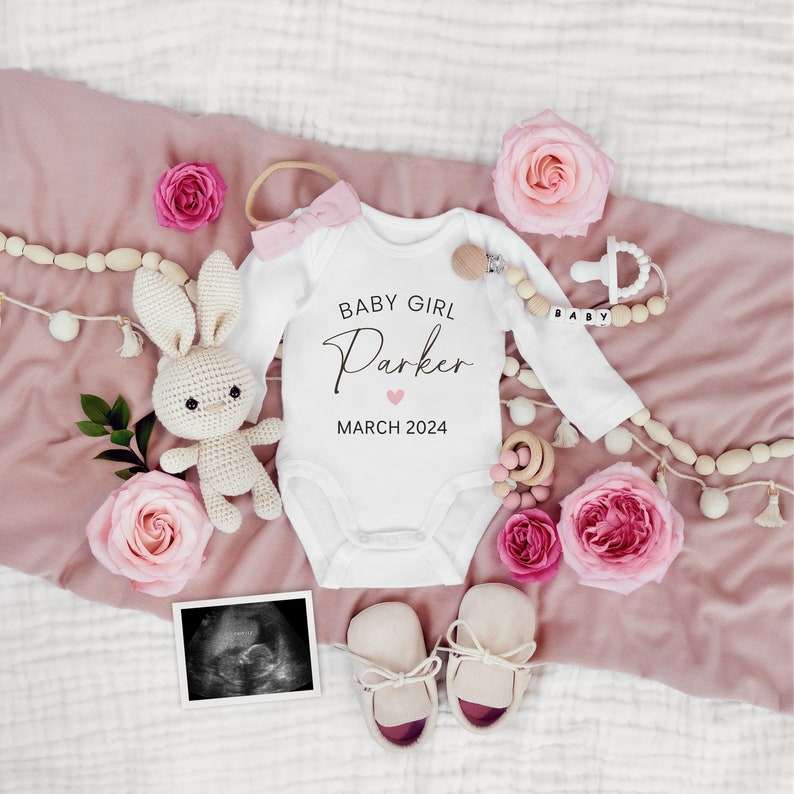 May include: A white onesie with the text "Baby Girl Parker March 2024" and a pink heart. The onesie is surrounded by pink roses, a crocheted bunny, a wooden rattle, and a pair of baby shoes.
