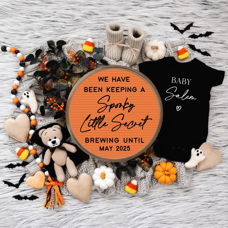 May include: A Halloween-themed baby announcement with a round orange sign that reads "We have been keeping a Spooky Little Secret Brewing Until May 2025".  A black onesie with the text "Baby Salem" is next to the sign.  The onesie is surrounded by Halloween decorations, including a teddy bear, candy corn, and bats.