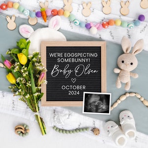 K&ouml;nnte beinhalten: Ein Schild aus Schiefer mit dem Text "We're eggspecting somebunny! Baby Olsen October 2024" umgeben von einer Vielzahl von Babyartikeln, darunter ein geh&auml;keltes H&auml;schen, ein Blumenstrau&szlig;, eine Girlande aus Holzhasen und ein Paar Babyschuhe.