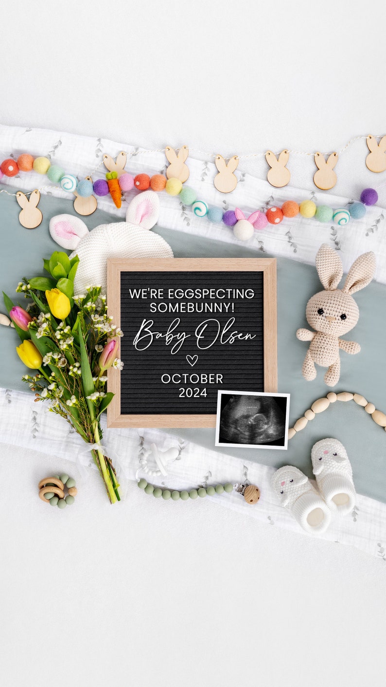 K&ouml;nnte beinhalten: Ein Schild aus Schiefer mit dem Text "We're eggspecting somebunny! Baby Olsen October 2024" umgeben von einer Vielzahl von Babyartikeln, darunter ein geh&auml;keltes H&auml;schen, ein Blumenstrau&szlig;, eine Girlande aus Holzhasen und ein Paar Babyschuhe.