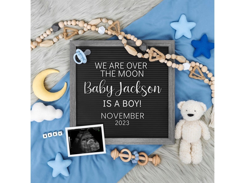 May include: A black letter board with the text "We are over the moon Baby Jackson is a boy! November 2023". The board is surrounded by a blue blanket, a yellow crescent moon, a white cloud, a pacifier, a wooden bead garland, a teddy bear, and a baby ultrasound image.