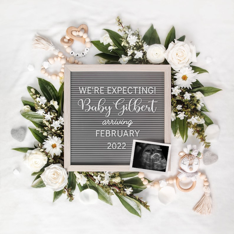 May include: A pregnancy announcement featuring a gray letter board with the text "WE'RE EXPECTING! Baby Gilbert arriving FEBRUARY 2022", surrounded by white flowers and greenery. An ultrasound image and baby toys are also present.