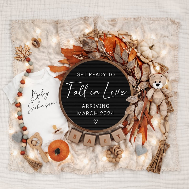 May include: A black letter board with the text "Get Ready To Fall in Love" and "Arriving March 2024" surrounded by autumnal decorations, including a white babygrow with the text "Baby Johnson", a small pumpkin, and wooden blocks spelling "BABY".