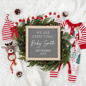 May include: A white letter board with the text "We are expecting! Baby Smith September 2023" surrounded by a green wreath with white lights. There is a red and white striped baby outfit, a red and white striped hat, a crocheted reindeer toy, and a red and white beaded garland.
