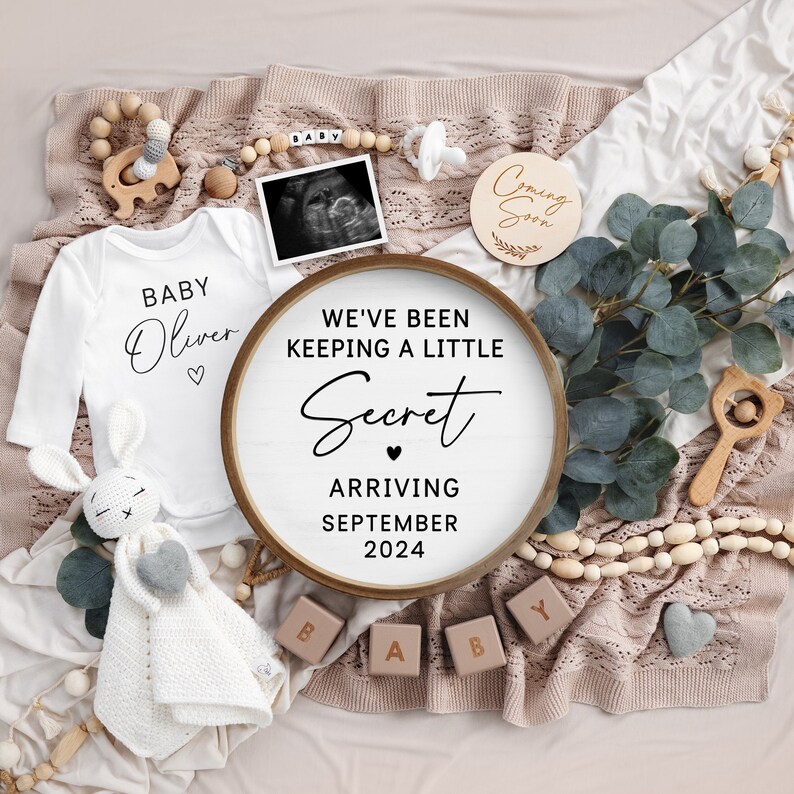 May include: A wooden sign with the text "We've been keeping a little secret" and "Arriving September 2024". The sign is surrounded by a white onesie with the text "Baby Oliver", a wooden elephant, a wooden rattle, a white crocheted bunny, a white crocheted blanket, a wooden block that says "Baby", and a garland of wooden beads.