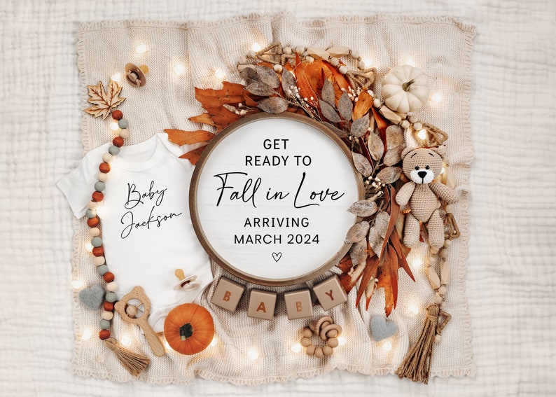 May include: A white babygrow with the text "Baby Jackson" in black script. A wooden sign with the text "Get Ready to Fall in Love Arriving March 2024" is surrounded by autumn-themed decorations, including a small teddy bear, a pumpkin, and a wreath of leaves.