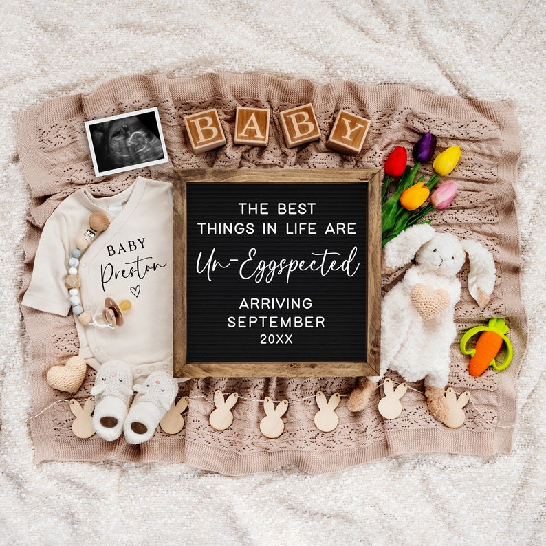 May include: A black letter board with the text "The best things in life are Un-Eggspected" and "Arriving September 20XX".  The board is surrounded by a white baby onesie with the text "Baby Preston", a white bunny rabbit toy, a pacifier, a pair of baby booties, and a string of wooden bunny rabbit cutouts.