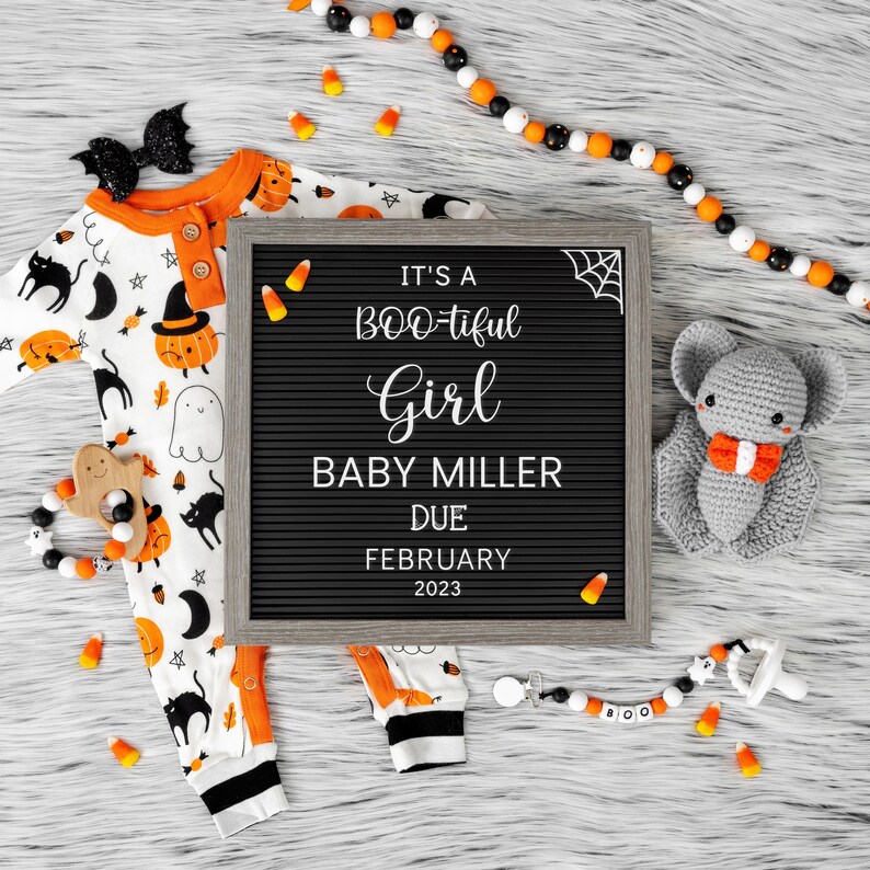 Puede incluir: Un tablero de letras negro con el texto "It's a BOO-tiful Girl Baby Miller Due February 2023". El tablero de letras est&aacute; rodeado de decoraciones de Halloween, incluyendo un mameluco para beb&eacute;, un murci&eacute;lago de crochet y ma&iacute;z dulce.