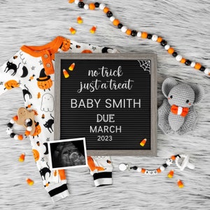 K&ouml;nnte beinhalten: Ein schwarzes Buchstabenbrett mit dem Text "no trick just a treat Baby Smith Due March 2023". Ein wei&szlig;er und orangefarbener Baby-Strampler mit einem schwarz-wei&szlig;en Halloween-Druck, eine graue geh&auml;kelte Fledermaus und ein schwarz-wei&szlig;er Schnuller mit dem Wort "BOO" darauf sind um das Buchstabenbrett herum angeordnet.
