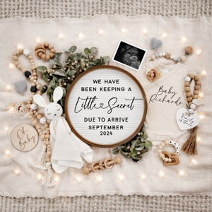 May include: A wooden sign with the text "We have been keeping a little secret due to arrive September 2024". The sign is surrounded by a variety of baby items, including a white onesie, a wooden rattle, a stuffed bunny, and a wooden bead necklace.