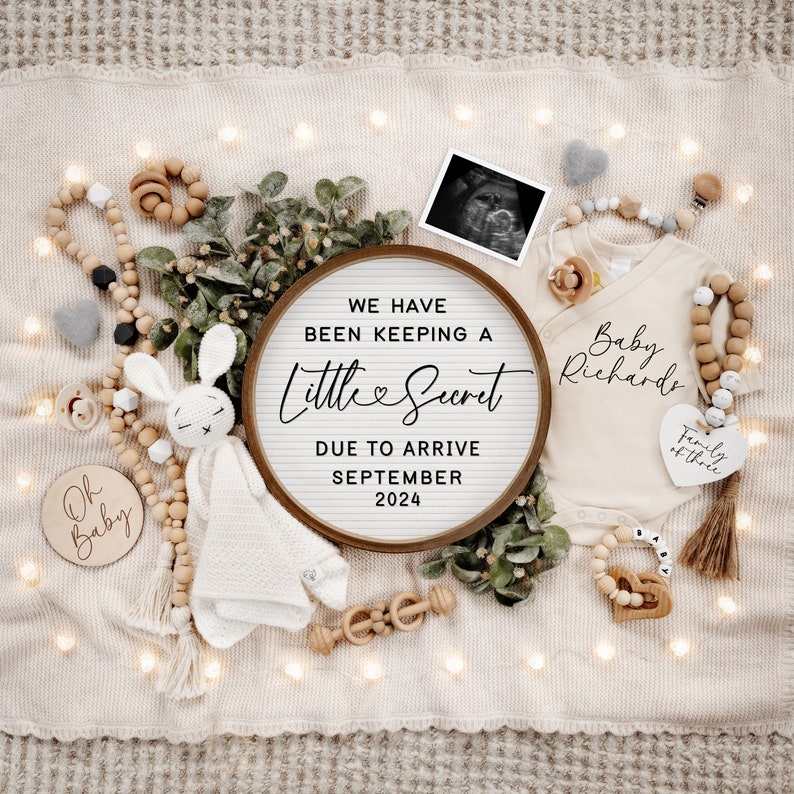 May include: A wooden sign with the text "We have been keeping a little secret due to arrive September 2024". The sign is surrounded by a variety of baby items, including a white onesie, a wooden rattle, a stuffed bunny, and a wooden bead necklace.