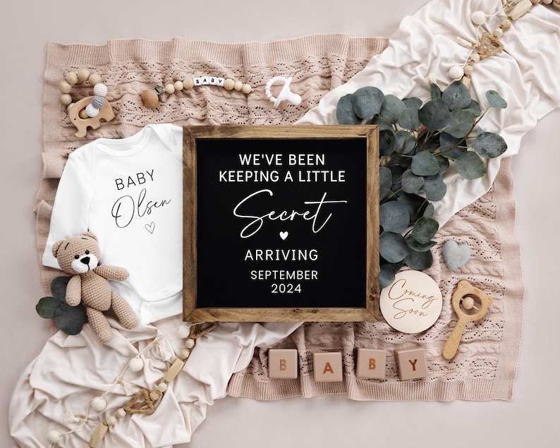 Op de afbeelding: Een baby-aankondiging met een wit rompertje met de tekst "Baby Olsen" en een zwart schoolbord met de tekst "We've been keeping a little secret arriving September 2024".