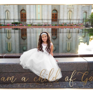 May include: A young girl in a white dress sits on a stone ledge in front of a large building with a reflecting pool in the foreground. The text "i am a child of God" is written in gold script on the ground in front of her.