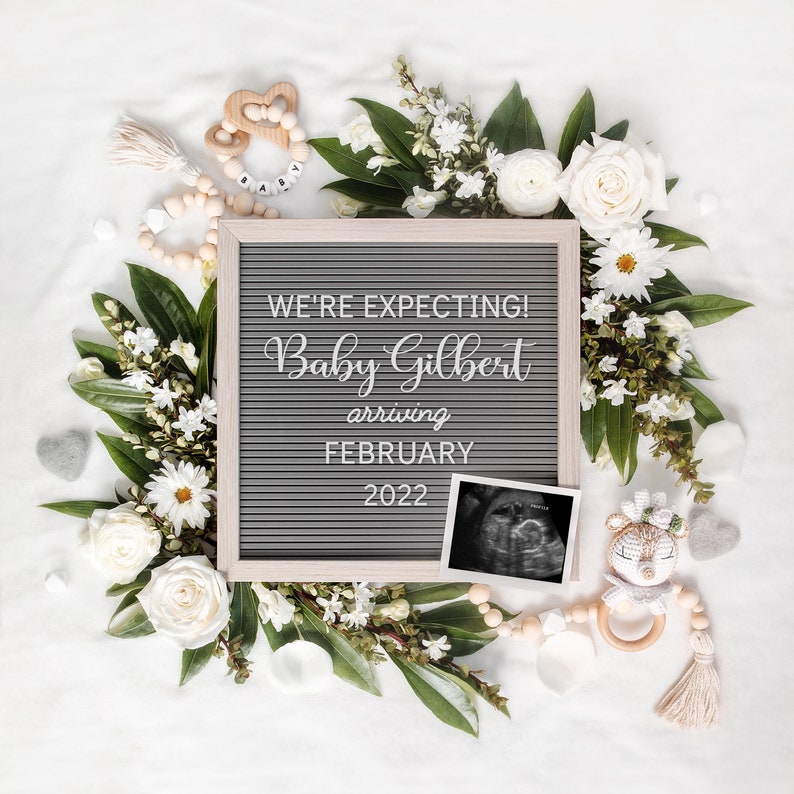 May include: A pregnancy announcement featuring a gray letter board in a light wood frame, surrounded by white flowers and foliage. The board states "WE'RE EXPECTING! Baby Gilbert arriving FEBRUARY 2022." An ultrasound scan and baby toys are also present.