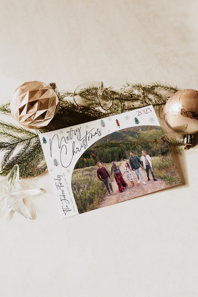 May include: A Christmas card with a photo of a family of five walking on a path in a forest. The card has a white background with green and red Christmas trees and snowflakes. The text "Merry Christmas" is written in a cursive font and the year "2025" is printed in the top right corner. The text "The Jackson Family" is printed in a black font on the left side of the card.