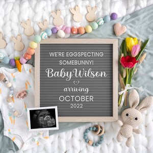 May include: A gray letter board with the text "We're eggspecting somebunny! Baby Wilson arriving October 2022". The board is surrounded by a garland of pastel colored felt balls and wooden bunny shapes. There is a bouquet of tulips, a crocheted bunny, a baby onesie, and a sonogram image.