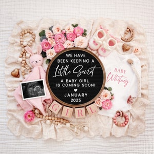 K&ouml;nnte beinhalten: Ein schwarzes Buchstabenbrett mit dem Text "We have been keeping a little secret. A baby girl is coming soon! January 2025."  Rosa und wei&szlig;e Babykleidung, eine rosa Hasenrassel und ein Ultraschallbild sind um das Buchstabenbrett herum angeordnet.