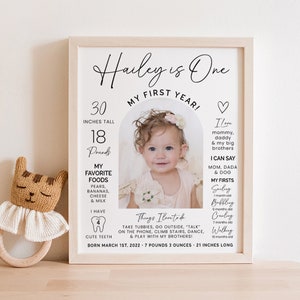 May include: A framed first birthday poster for a baby girl named Hailey. The poster features a photo of the baby, her height and weight, favorite foods, and things she loves to do. The text reads "Hailey is One, My First Year!" and includes the date of her birth: March 1st, 2022.
