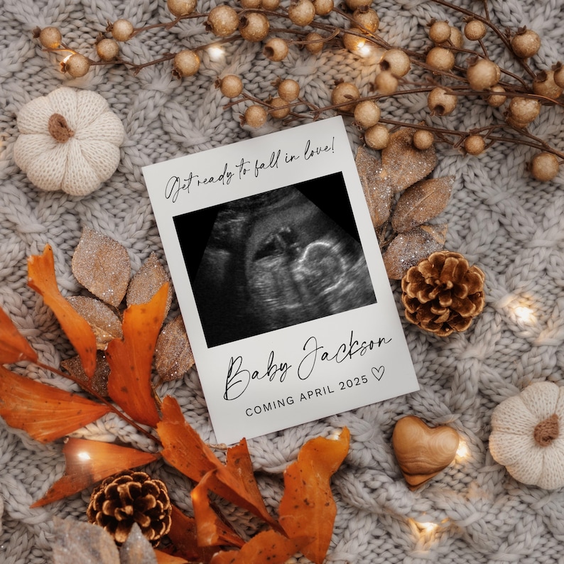 K&ouml;nnte beinhalten: Eine wei&szlig;e Karte mit einem Schwarzwei&szlig;-Ultraschallbild und dem Text "Get ready to fall in love!" Der Text "Baby Jackson" und "Coming April 2025" ist in schwarzer Kursivschrift geschrieben. Die Karte ist von Herbstdekorationen umgeben, darunter Tannenzapfen, Bl&auml;tter und ein wei&szlig;er K&uuml;rbis.