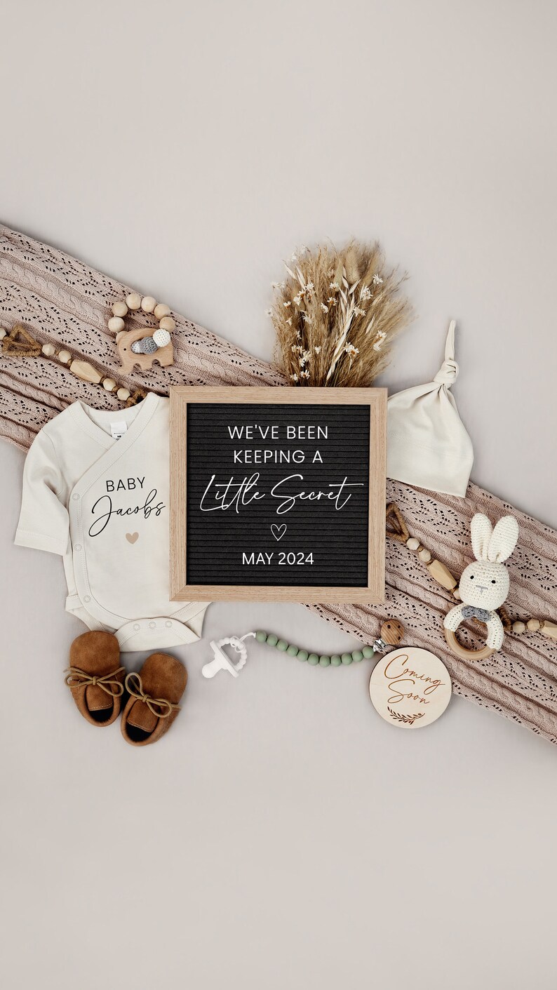 Puede incluir: Un anuncio de nacimiento con un body blanco que dice "Baby Jacobs" y un tablero de letras que dice "We've been keeping a little secret May 2024". Tambi&eacute;n hay zapatos de beb&eacute;, un sonajero de madera, un conejito de madera y un letrero de madera que dice "Coming Soon".
