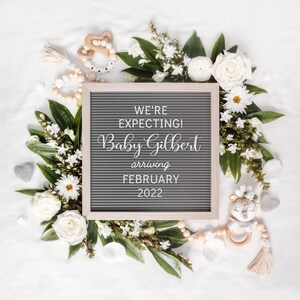 May include: A grey letter board with the text "WE'RE EXPECTING! Baby Gilbert arriving FEBRUARY 2022", surrounded by white flowers, greenery, and baby toys. The arrangement is on a white background, ideal for a pregnancy announcement.