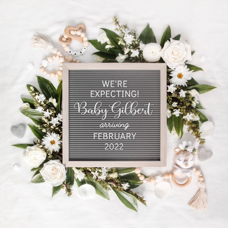May include: A grey letter board with the text "WE'RE EXPECTING! Baby Gilbert arriving FEBRUARY 2022", surrounded by white flowers, greenery, and baby toys. The arrangement is on a white background, ideal for a pregnancy announcement.