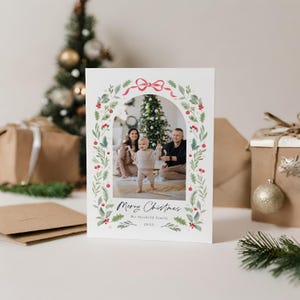 May include: A Christmas card with a watercolor wreath design featuring a red bow and green foliage. A family photo is inside the wreath, showing a couple and their baby standing in front of a Christmas tree. The card reads "Merry Christmas The Mansfield Family 20XX".