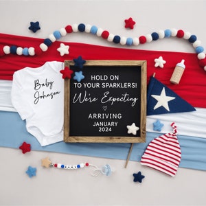 Puede incluir: Un letrero de pizarra con el texto "Hold on to your sparklers! We're expecting. Arriving January 2024."  El letrero est&aacute; rodeado de art&iacute;culos de beb&eacute; rojo, blanco y azul, incluyendo un body, un gorro y un sonajero.