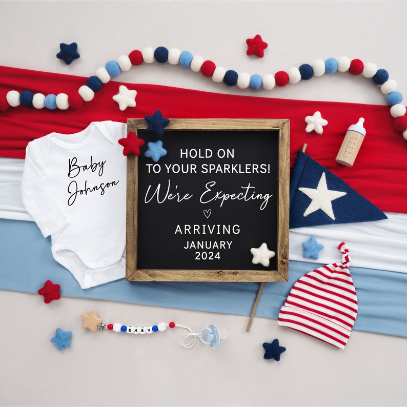 Puede incluir: Un letrero de pizarra con el texto "Hold on to your sparklers! We're expecting. Arriving January 2024."  El letrero est&aacute; rodeado de art&iacute;culos de beb&eacute; rojo, blanco y azul, incluyendo un body, un gorro y un sonajero.