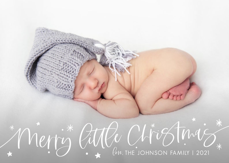 May include: A newborn baby wearing a gray knitted hat with a white tassel, sleeping on a white blanket. The text "Merry little Christmas" is written in white cursive script below the baby, with the words "love, THE JOHNSON FAMILY | 2021" written below the text.