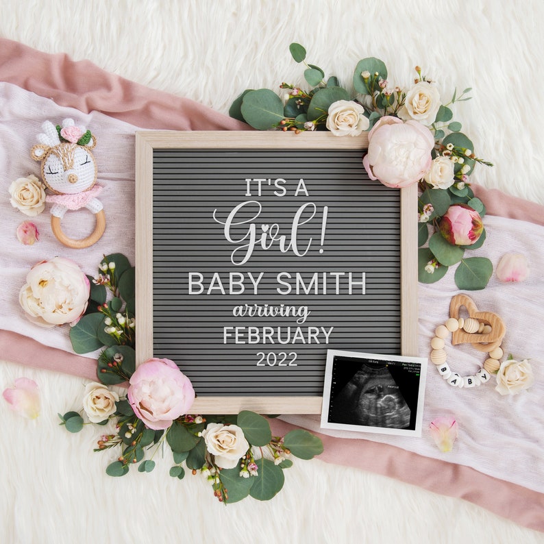Może przedstawiać: Szara tablica z literami z tekstem "It's a Girl! Baby Smith arriving February 2022". Tablica jest otoczona r&oacute;żowymi i białymi kwiatami, grzechotką dla dziecka i obrazem USG.