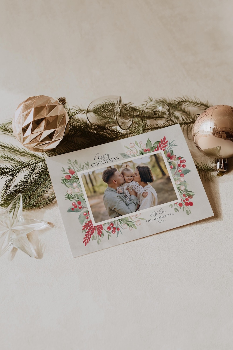 Pode incluir: Um cart&atilde;o de Natal com uma borda de aquarela de bagas vermelhas e folhas verdes. Uma foto de uma fam&iacute;lia de tr&ecirc;s pessoas est&aacute; no centro do cart&atilde;o. O texto "Merry Christmas" est&aacute; no topo do cart&atilde;o e "with love, the Hamiltons - 2024" est&aacute; na parte inferior.