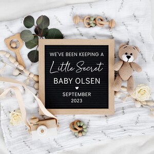 K&ouml;nnte beinhalten: Ein schwarzes Buchstabenbrett mit wei&szlig;em Text, der "We've been keeping a little secret, Baby Olsen, September 2023" ank&uuml;ndigt. Das Brett ist von einer wei&szlig;en Decke, einem Teddyb&auml;r, Holzspielzeug und Gr&uuml;n umgeben.