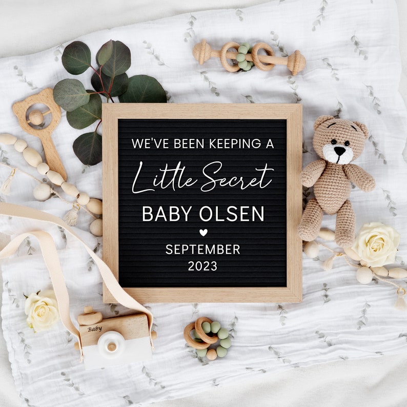 K&ouml;nnte beinhalten: Ein schwarzes Buchstabenbrett mit wei&szlig;em Text, der "We've been keeping a little secret, Baby Olsen, September 2023" ank&uuml;ndigt. Das Brett ist von einer wei&szlig;en Decke, einem Teddyb&auml;r, Holzspielzeug und Gr&uuml;n umgeben.