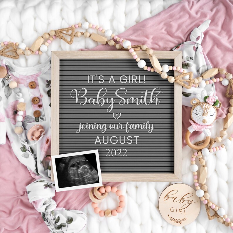 Może przedstawiać: Szara tablica z literami z tekstem "It's a girl! Baby Smith joining our family August 2022". Jest zdjęcie USG w czerni i bieli, r&oacute;żowo-biały str&oacute;j dla dziecka, drewniana grzechotka dla dziecka i drewniany znak z napisem "baby girl" na r&oacute;żowo-białym kocu.