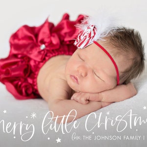 May include: A newborn baby girl wearing a red satin outfit with a white bow and a red and white candy cane bow on her head. The baby is sleeping with her head resting on her arm. The text "Merry little Christmas" is written in white cursive script below the baby. The text "love, THE JOHNSON FAMILY | 2021" is written in white below the cursive text.